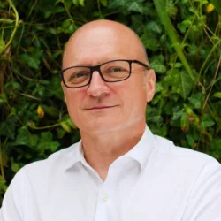 Slawomir standing in front of a camera wearing white shirt with plants on the background.