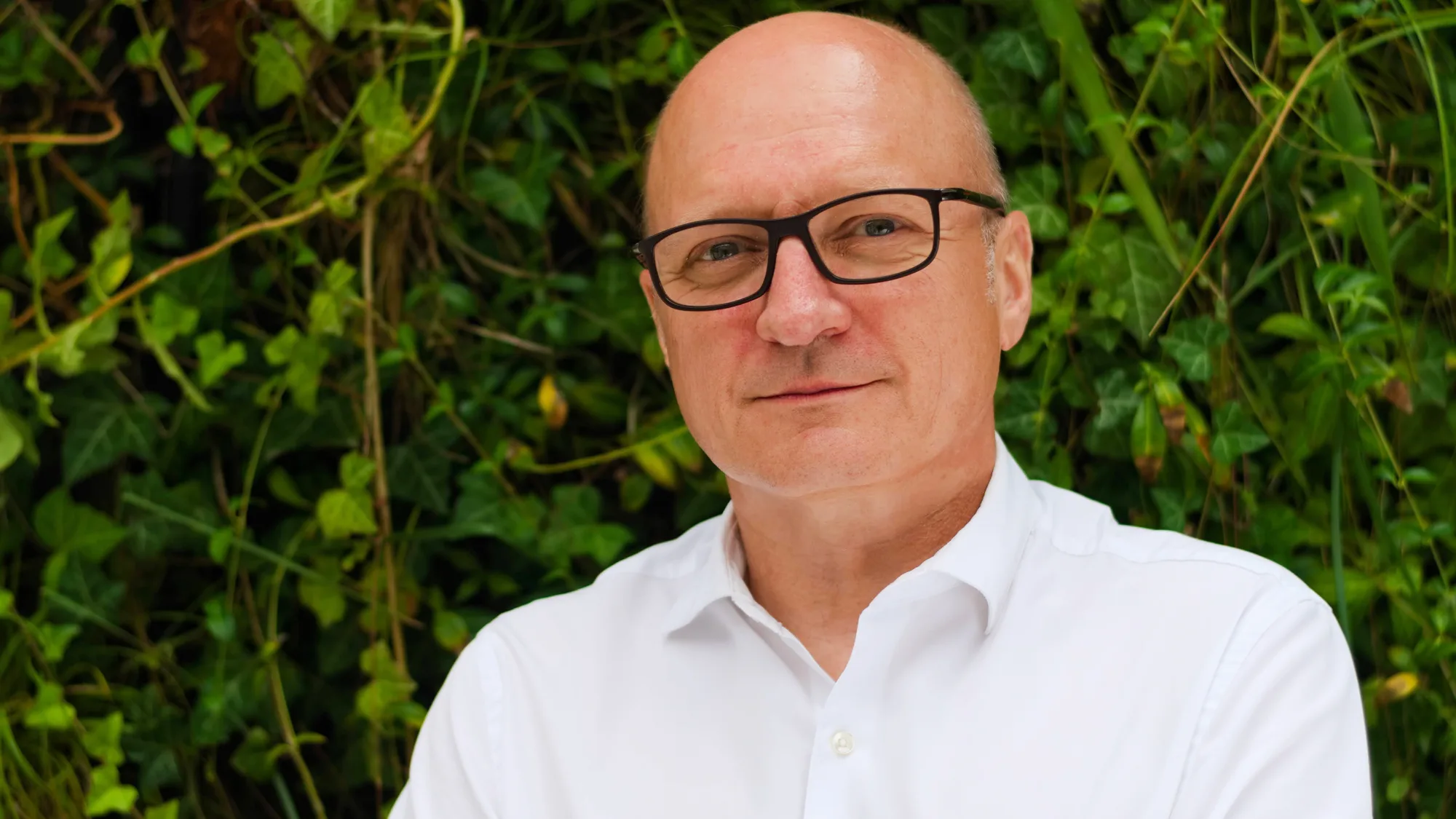 Slawomir standing in front of a camera wearing white shirt with plants on the background.