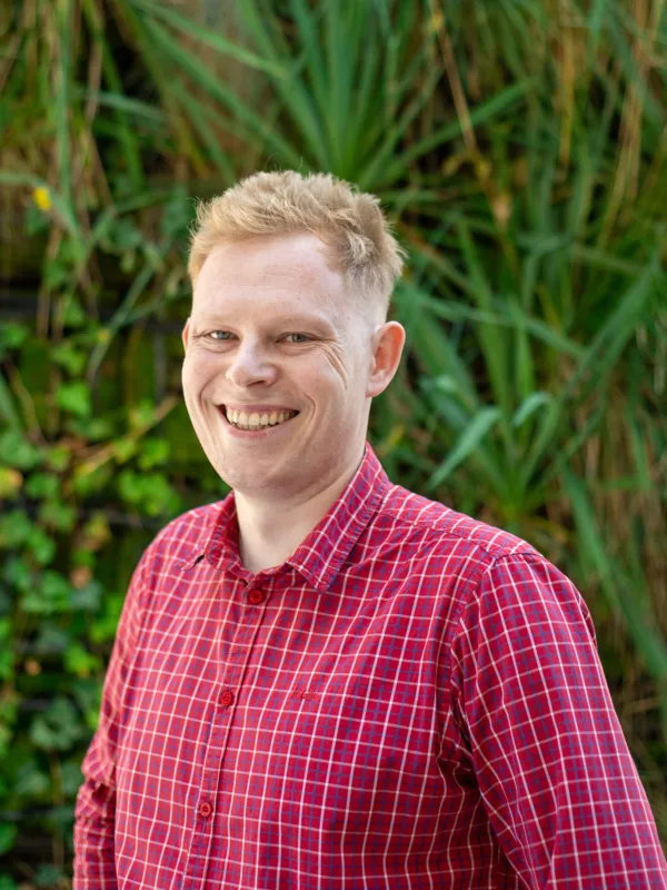 Przemyslaw in a red check shirt smiling to camera outside in front of a green hedge