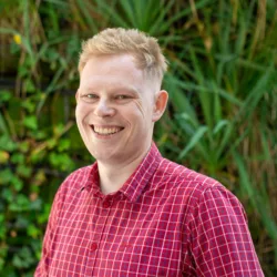 Przemyslaw in a red check shirt smiling to camera outside in front of a green hedge