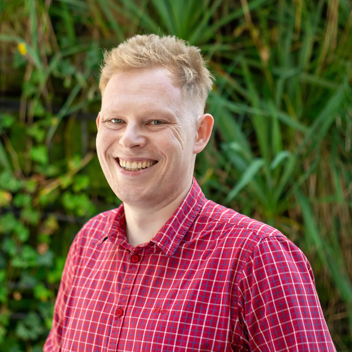 Przemyslaw in a red check shirt smiling to camera outside in front of a green hedge