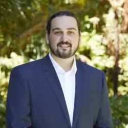 Head and sholders shot of Stefan wearing white shirt with blue blazer against a background of foliage