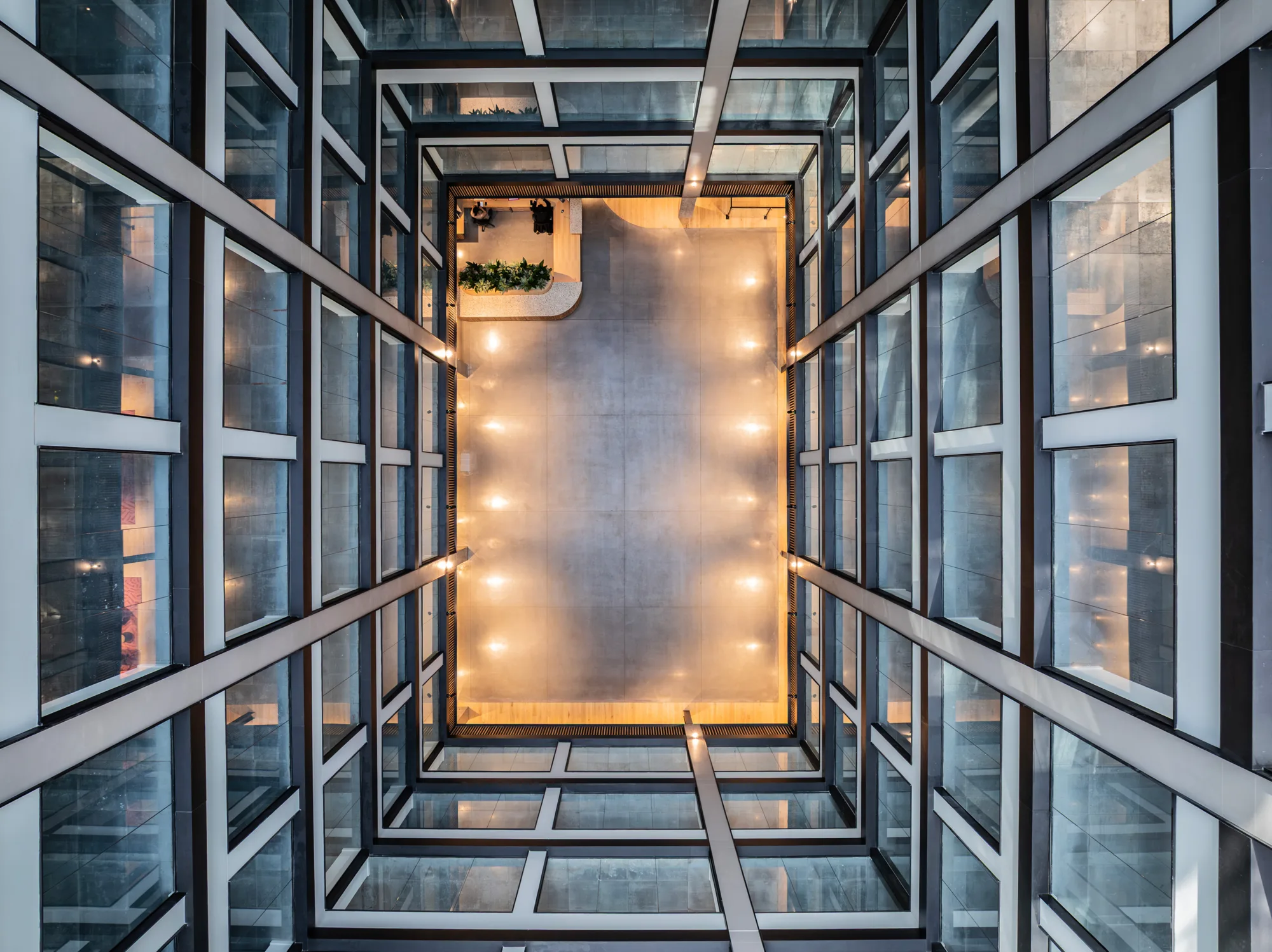 View looking downward through a multi-storey glass atrium with black-framed windows and illuminated walkways surrounding a central open space. A curved reception desk with a planter is visible on the ground floor. Two small, indistinct people appear near the desk.