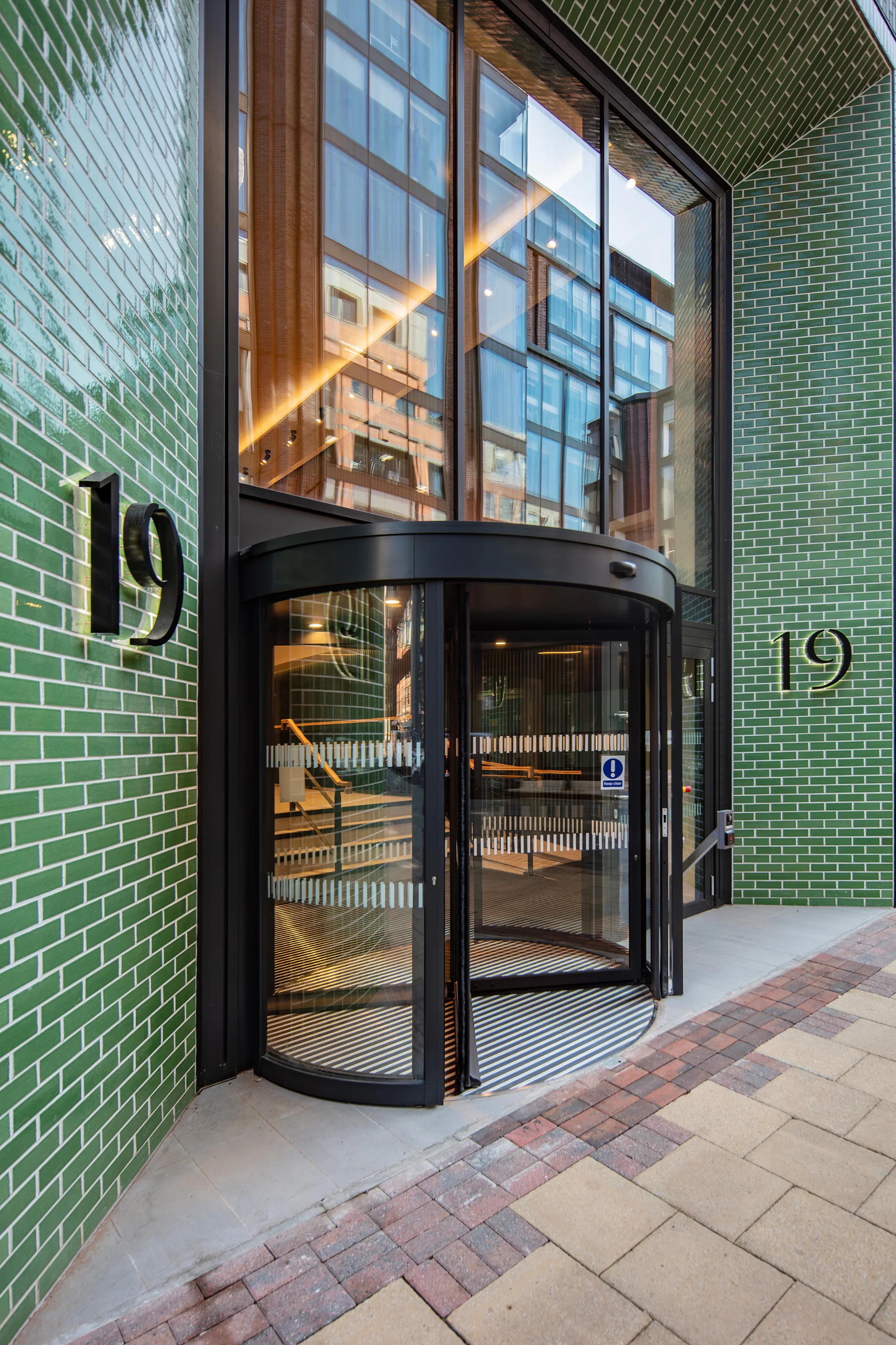 Exterior entrance of a building clad in green glazed brick tiles, featuring a black-framed revolving glass door. The number “19” appears on both sides of the entrance. Reflections of nearby buildings are visible in the tall glass façade, with a paved sidewalk in the foreground and no people present.
