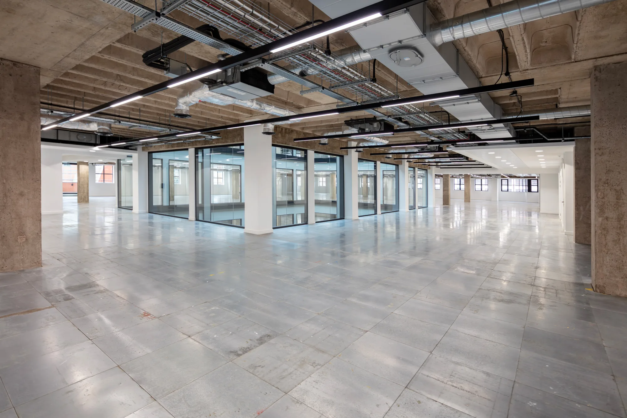 Open-plan office interior with polished gray flooring, exposed concrete ceilings, visible ductwork and cable trays, glass-walled rooms, and structural columns. Linear suspended lights run across the ceiling.