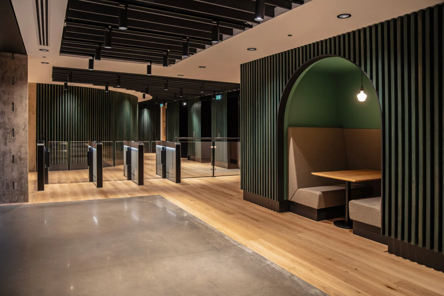 Modern office interior with wood flooring, dark slatted wall panels, and ceiling-mounted lighting. Glass access turnstiles line a hallway, while an arched booth with bench seating, a table, and a hanging light occupies the right side.