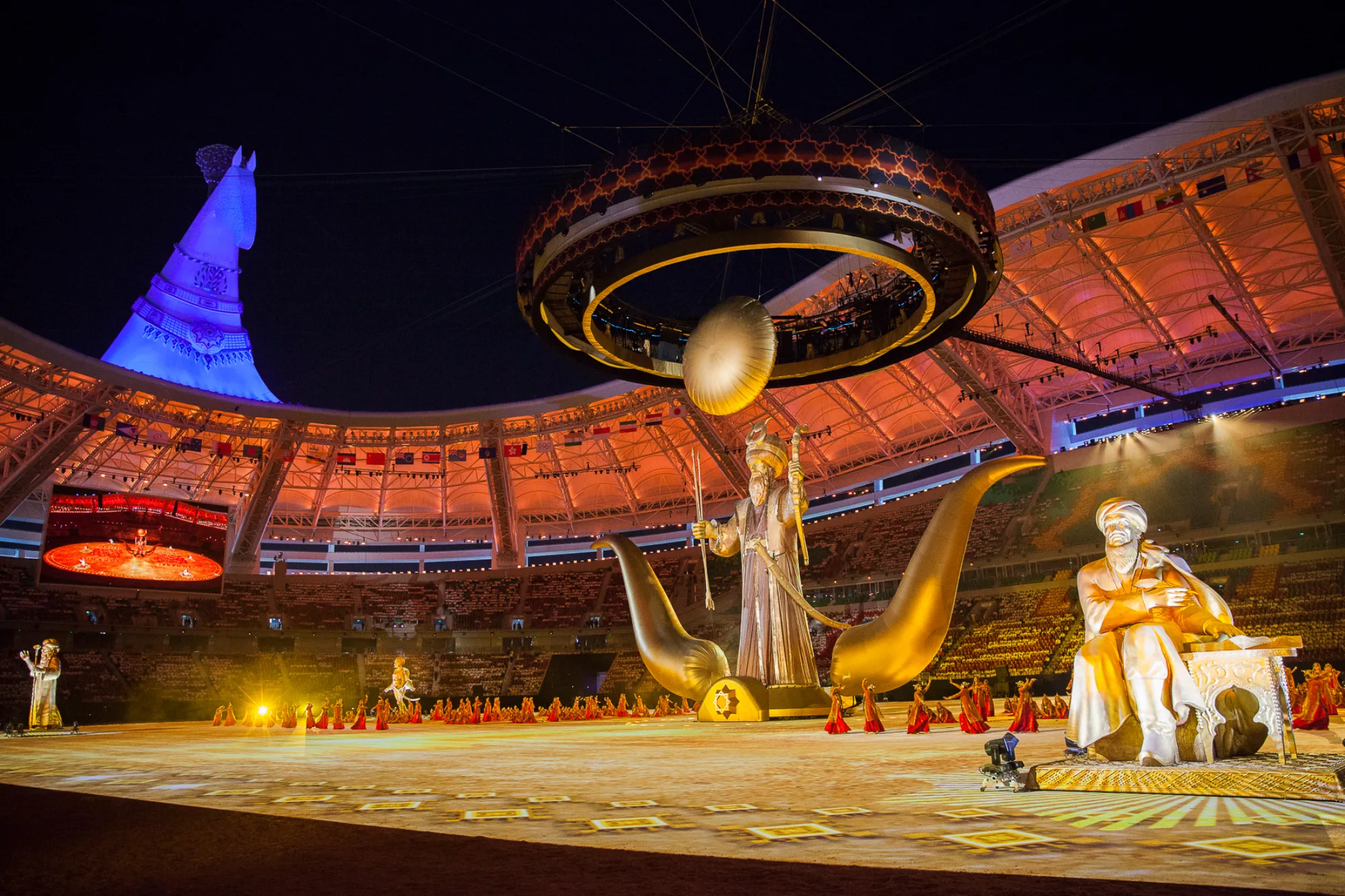 Four large sculptural figures on the main arena stage with dancers dressed in red