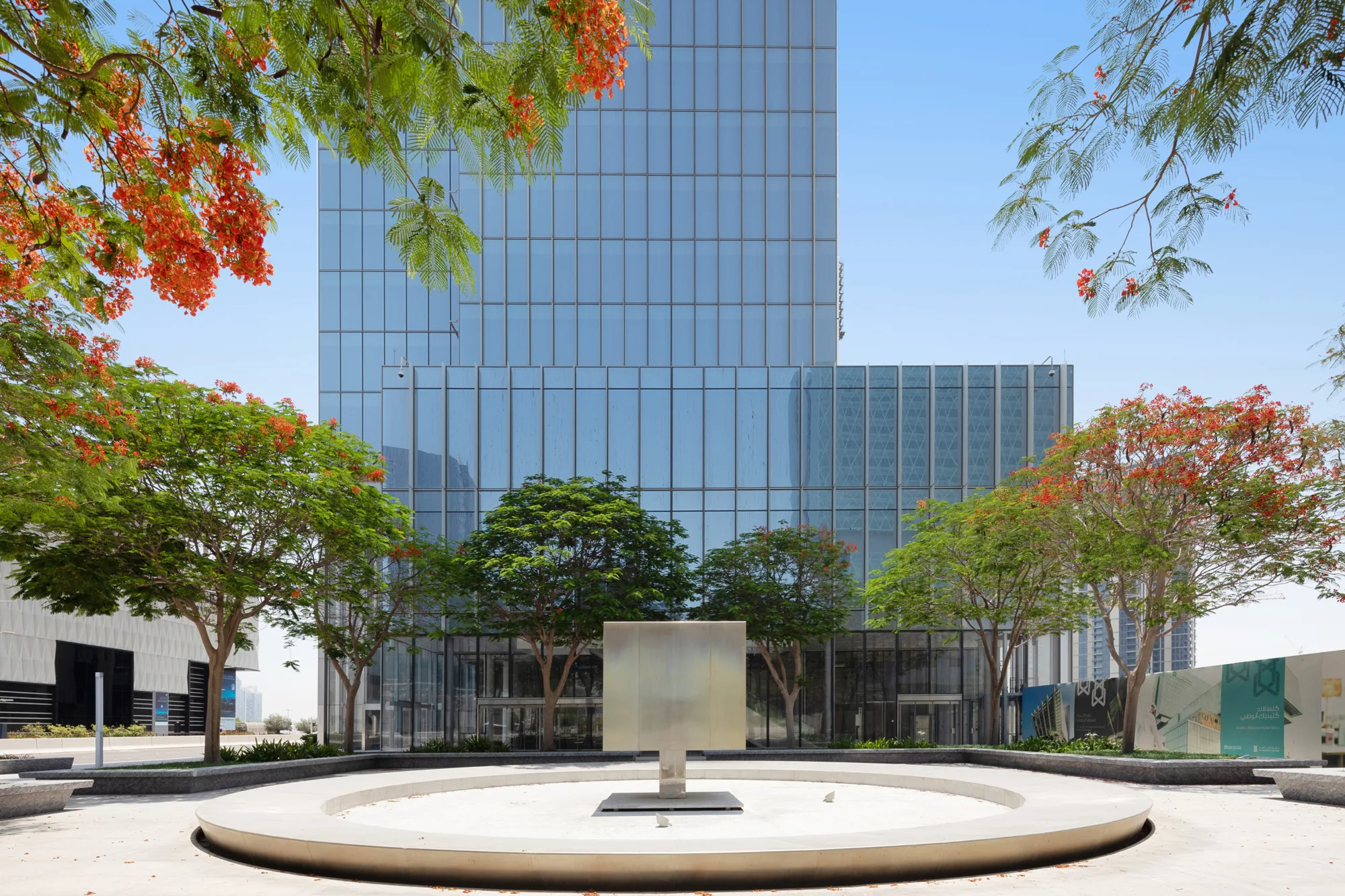 Front view of a modern high-rise building with a glass facade; landscaped area with trees and a central sculpture in foreground.