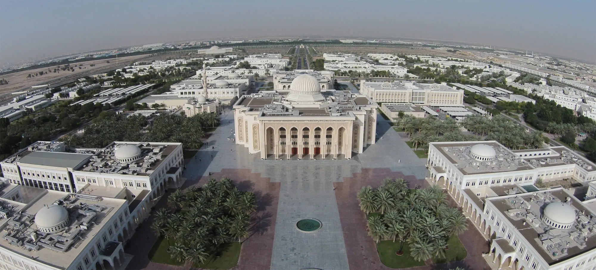 Aerial view of a large architectural complex with a central domed building featuring columns and arches, surrounded by smaller domed structures, landscaped areas with palm trees, paved walkways, and a circular fountain in the foreground.