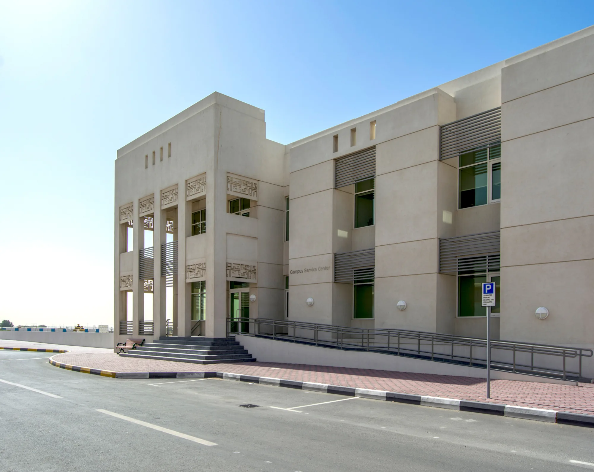 Modern institutional building with light beige walls, rectangular windows with metal slats, and a columned entrance featuring decorative panels; ramp access and marked parking spaces visible under a clear blue sky.