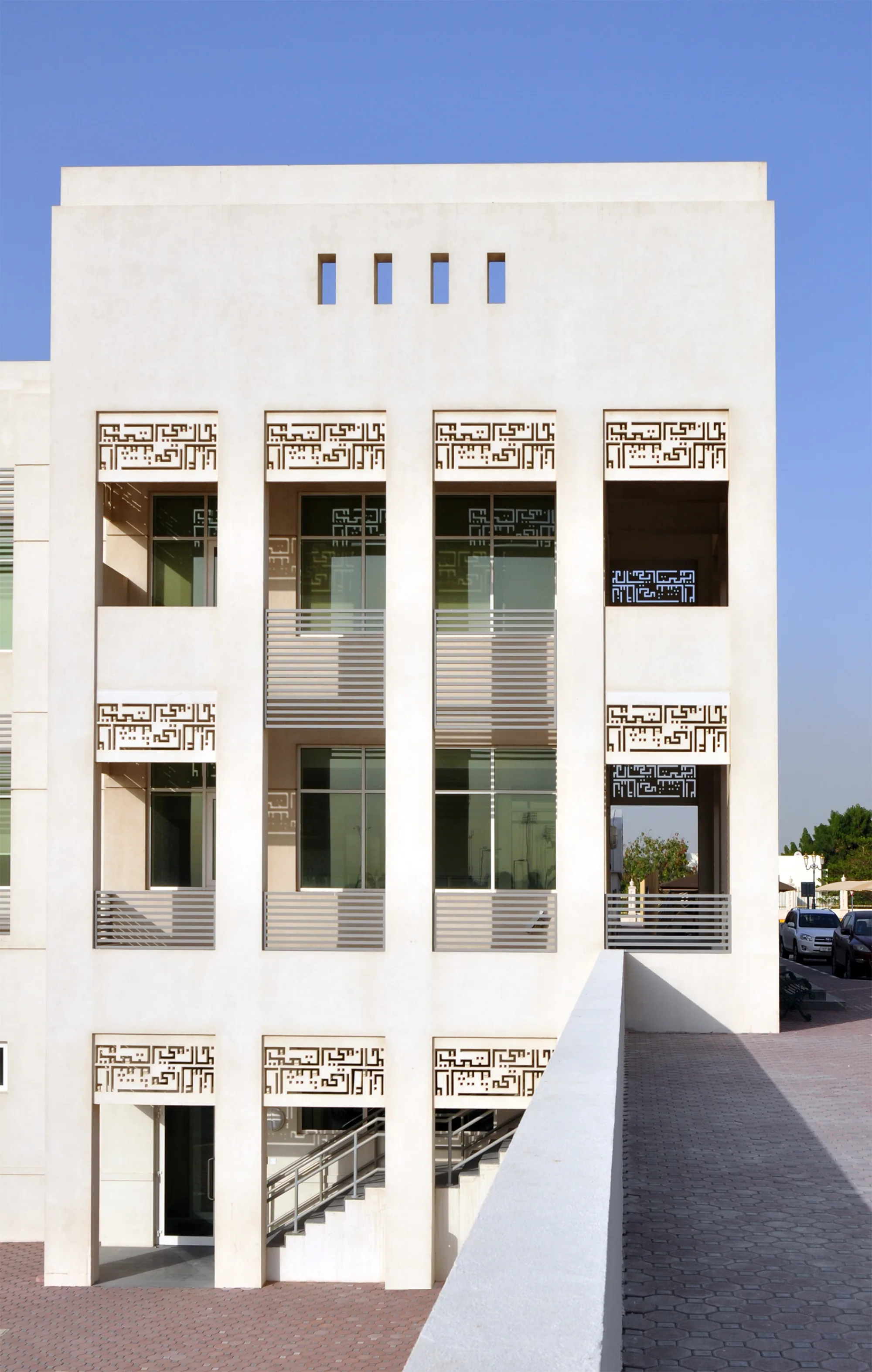 Modern white building with clean rectangular design, three vertical columns of windows framed by decorative geometric patterns, metal railings, and an entrance with stairs; paved area and parked vehicles visible under a clear blue sky.