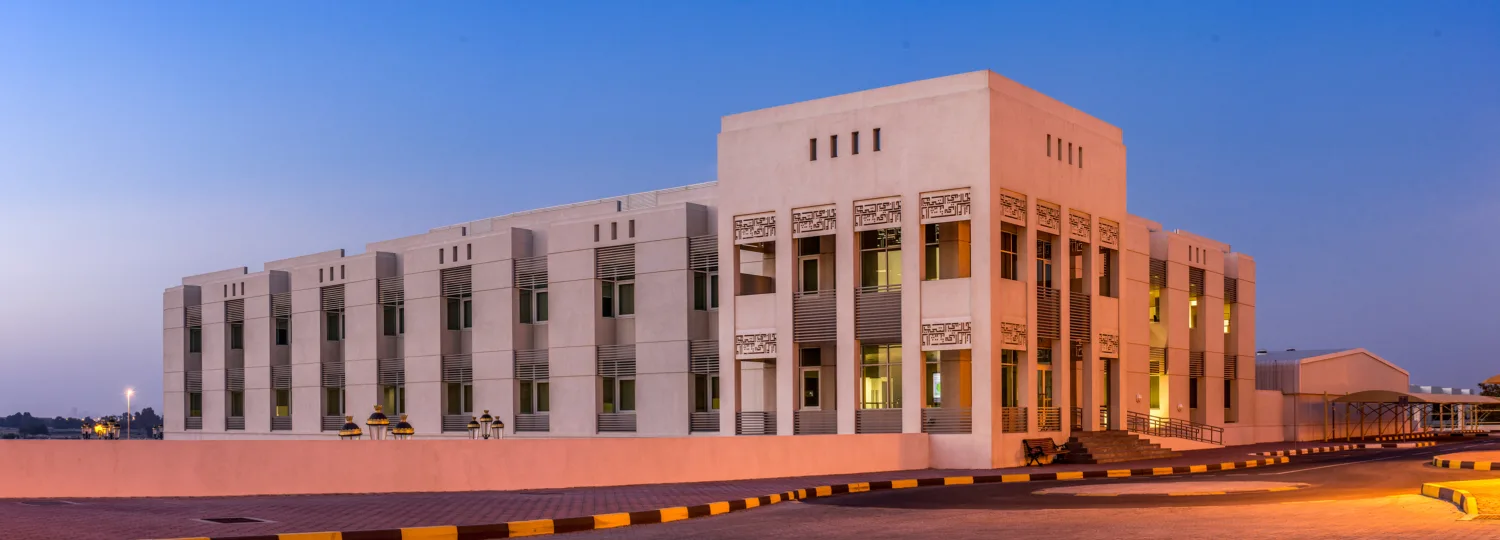 Modern rectangular building with light-coloured facade, large rows of windows, and decorative panels above some windows; curved road with black and yellow curbs in foreground under a clear twilight sky.