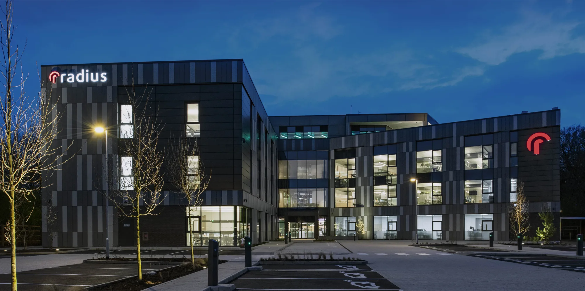 Image taken from a car park, with the camera pointing towards the radius office building lit brightly with a clear evening sky behind it