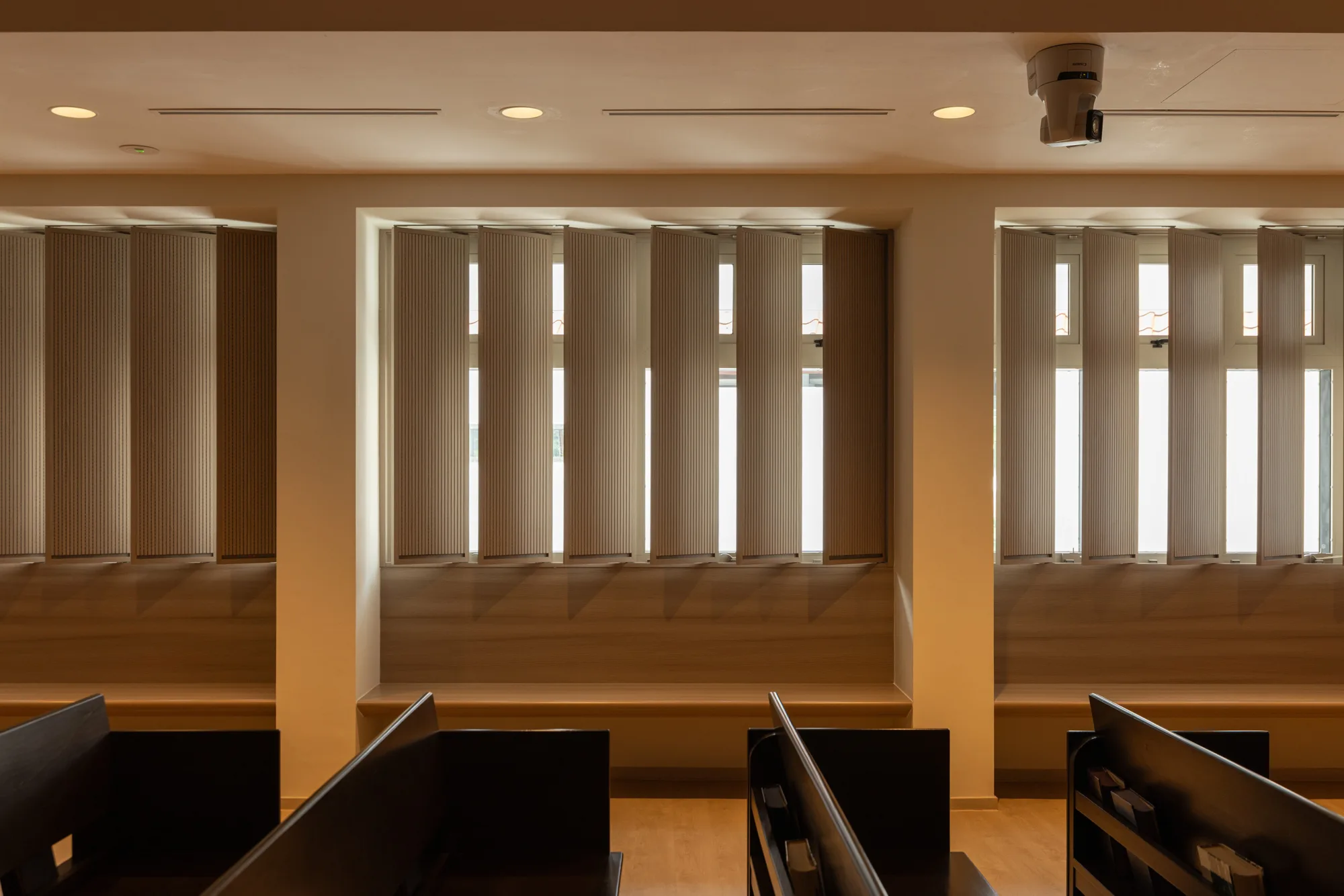Interior view of a room with three large windows covered by vertical blinds, beige walls, ceiling lights, and a mounted projector. Dark wooden pews are partially visible in the foreground.