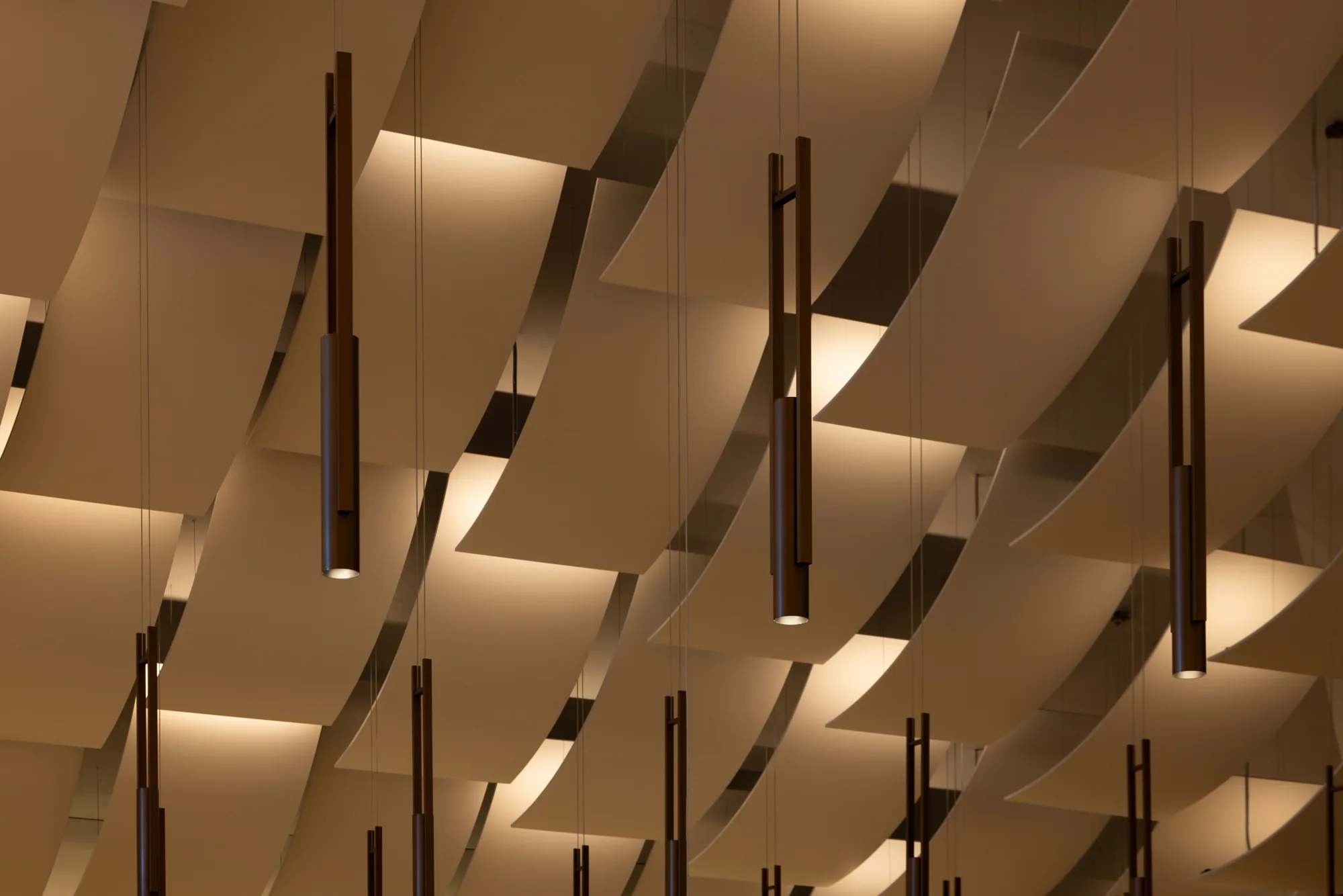 Close-up view of a ceiling with multiple curved, overlapping white panels arranged in rows. Several slim, cylindrical pendant lights hang vertically from the ceiling on thin cables.