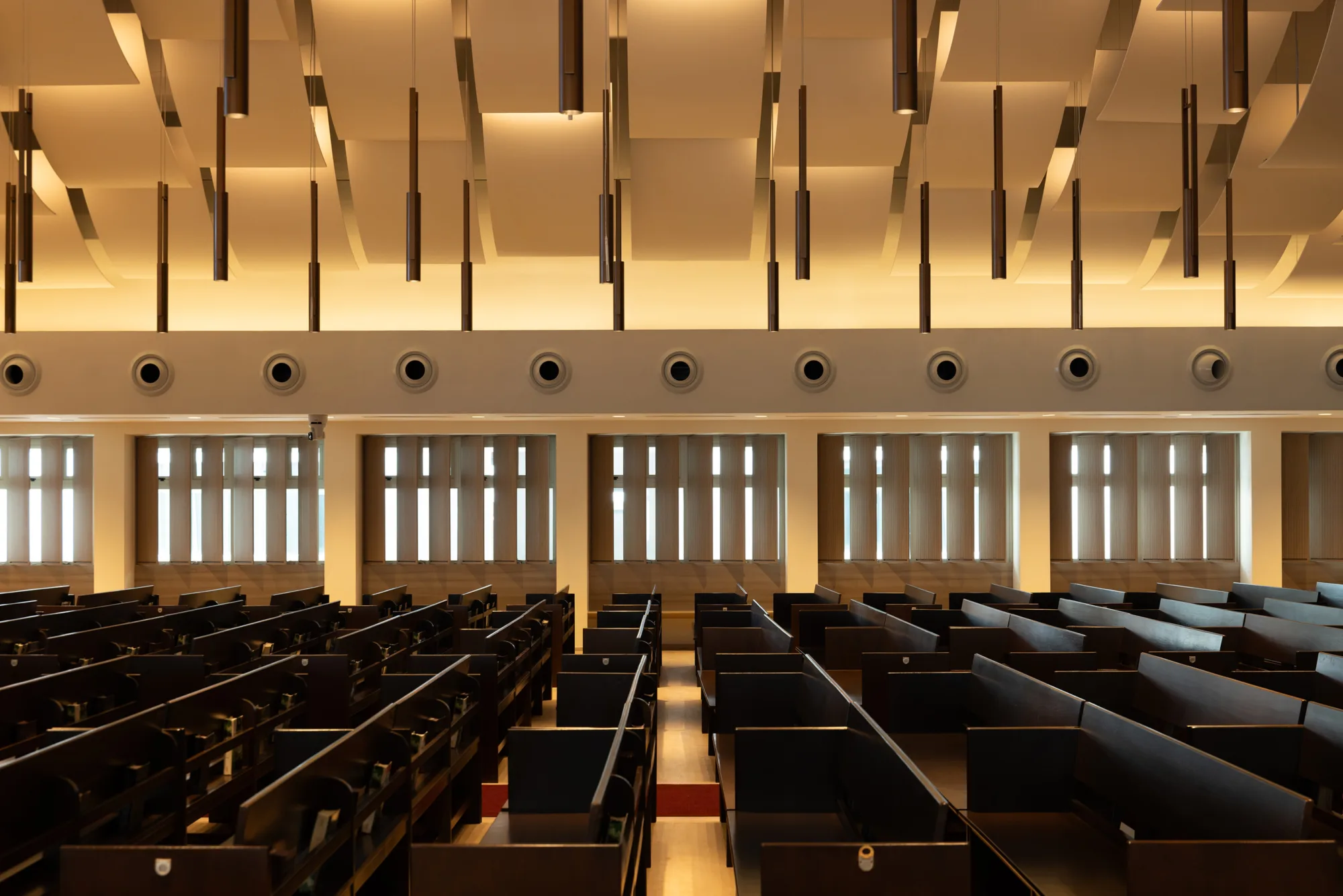 Rows of dark wooden pews arranged in parallel lines inside a large hall. The ceiling features suspended rectangular panels and cylindrical pendant lights. The back wall has multiple windows with vertical blinds and circular vents above them.