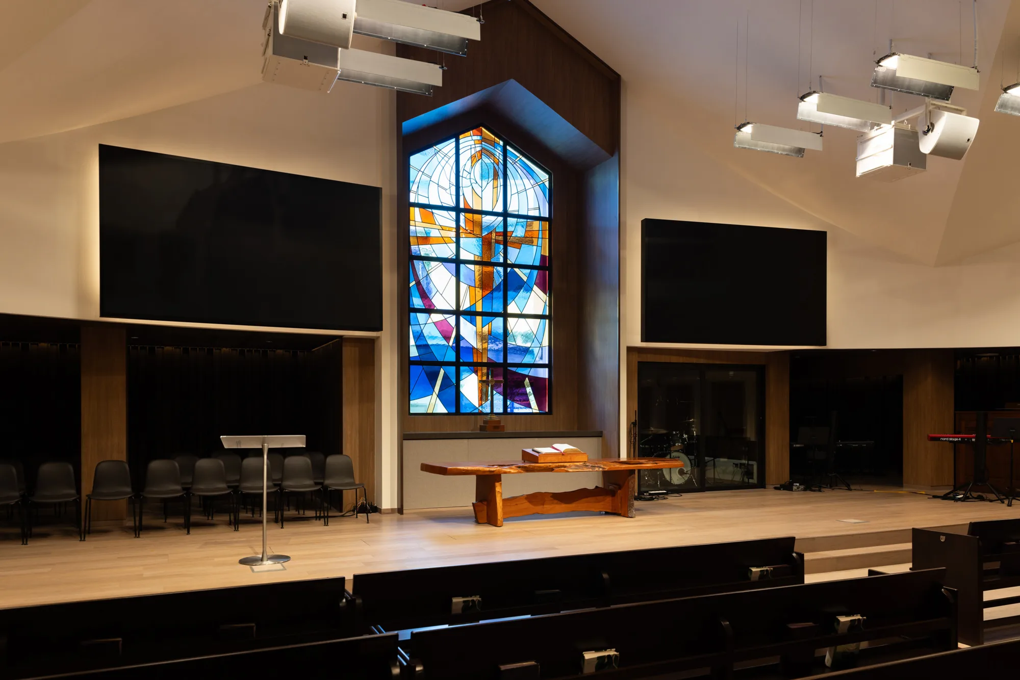 Stage area inside a church with a wooden altar table and an open book on top. Behind the stage is a large stained-glass window with geometric patterns in blue, orange, and white. Two large black screens are mounted on the wall, with chairs arranged along the back and ceiling lights above.