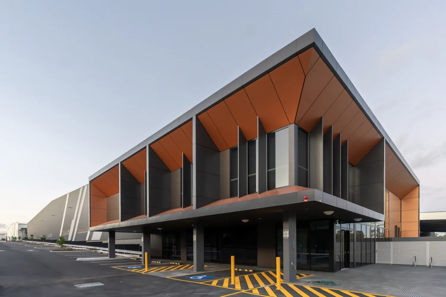 Modern commercial building with a cantilevered upper level, dark grey columns, and angled orange soffits. Glass façades line the ground floor, with marked parking bays and bollards in the foreground.