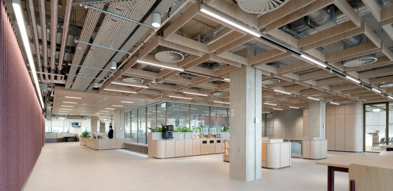 Modern open-plan office interior with exposed ceiling design, suspended lighting, glass-walled meeting rooms, light flooring, and modular counters with plants in a collaborative workspace.