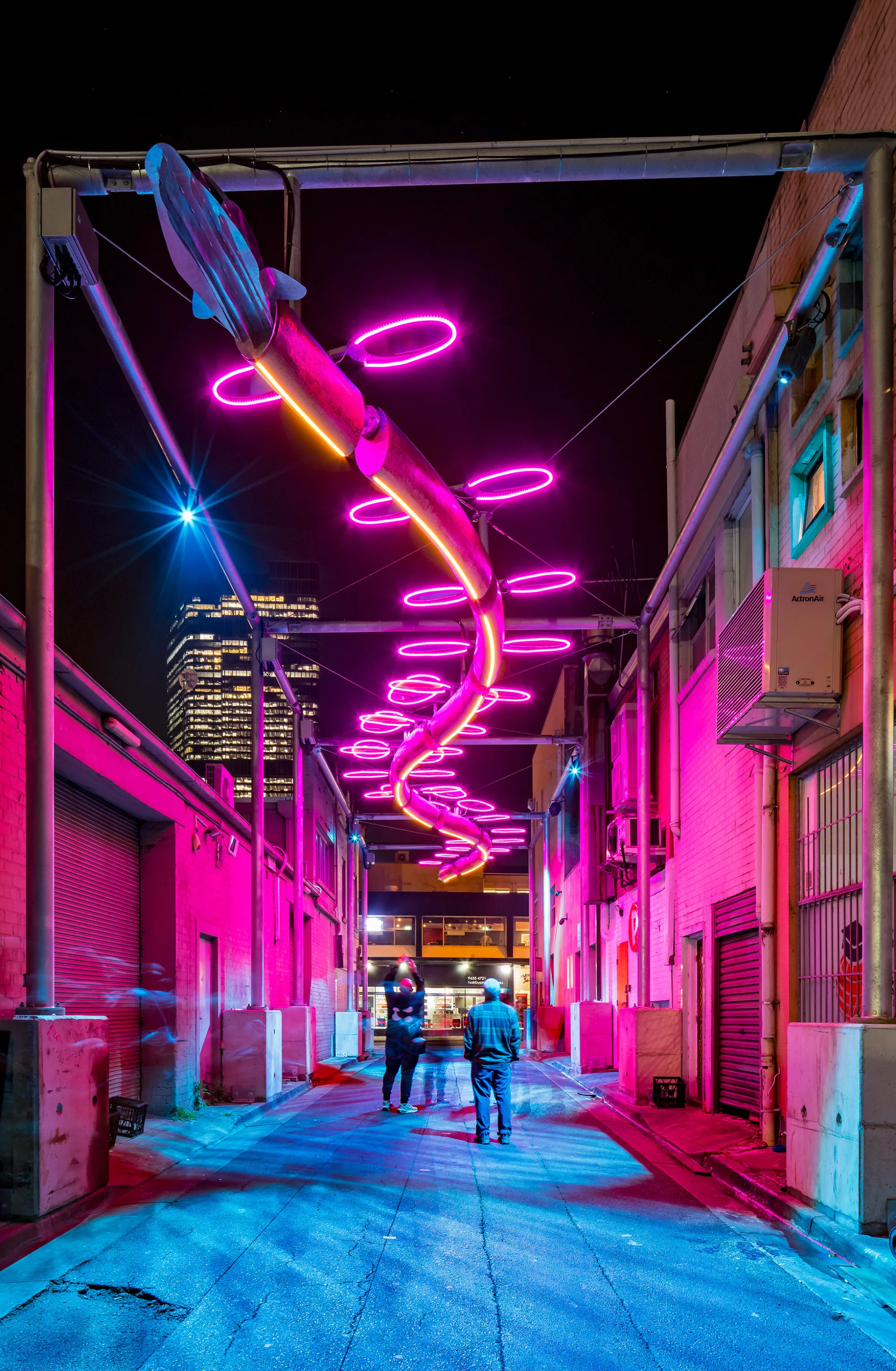 Nighttime alleyway featuring a twisting metallic structure with neon lights in purple, blue, green, and yellow forming circular patterns; buildings with windows and air conditioning units line both sides; tall lit buildings in background.