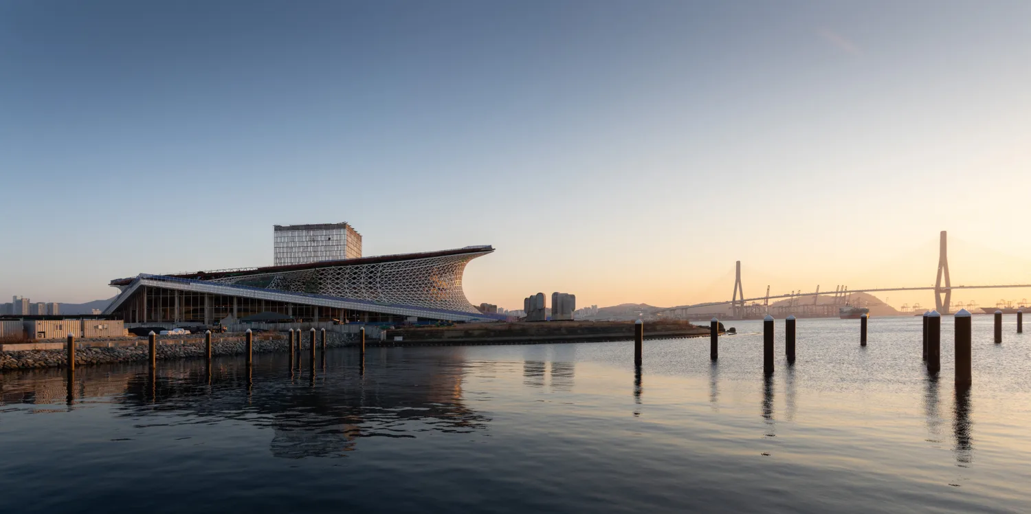 A wide-angle view of the Busan Opera House under construction on a calm bay, featuring the Busan Harbor Bridge.