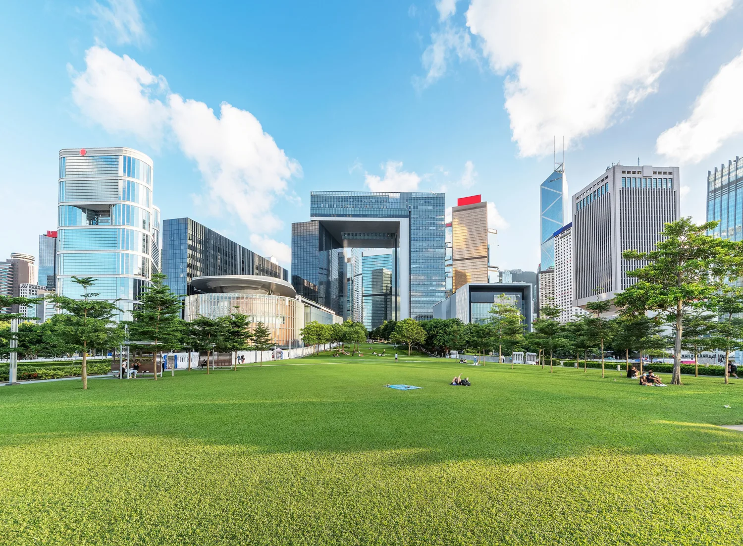 A wide green lawn in the foreground with scattered people, bordered by trees and modern high-rise buildings in the background. The skyline includes glass and steel structures under a bright blue sky with white clouds.
