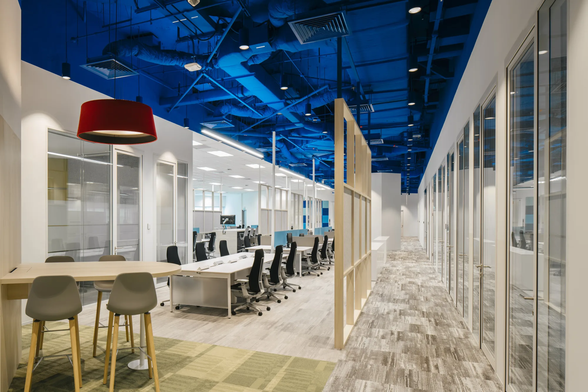 Carpeted workspace featuring row of individual desks facing each other and a table with high-top chairs on one side; flanked by hallways with glass doors along each wall