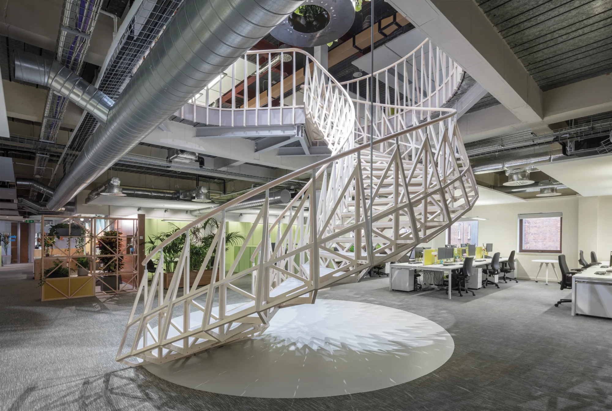 Side view of a white spiral staircase with a geometric pattern, in the centre of office desks and chairs