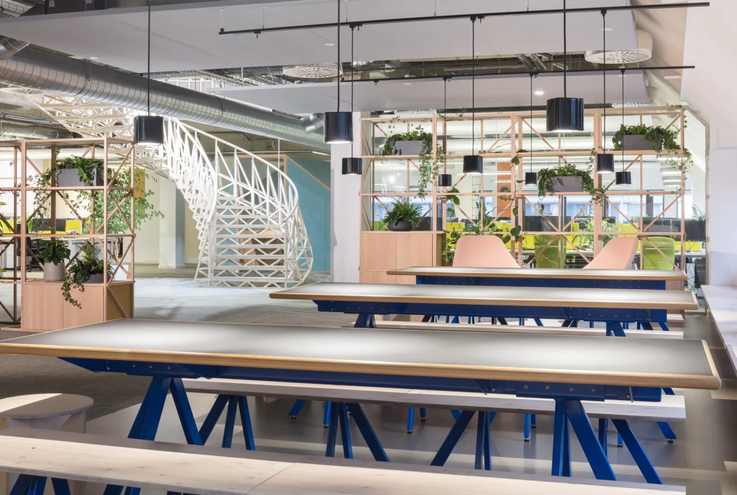 Interior of Cundall Birmingham office, grey topped dining tables int he foreground with a white spiral staircase and plants on wooden shelves in the background