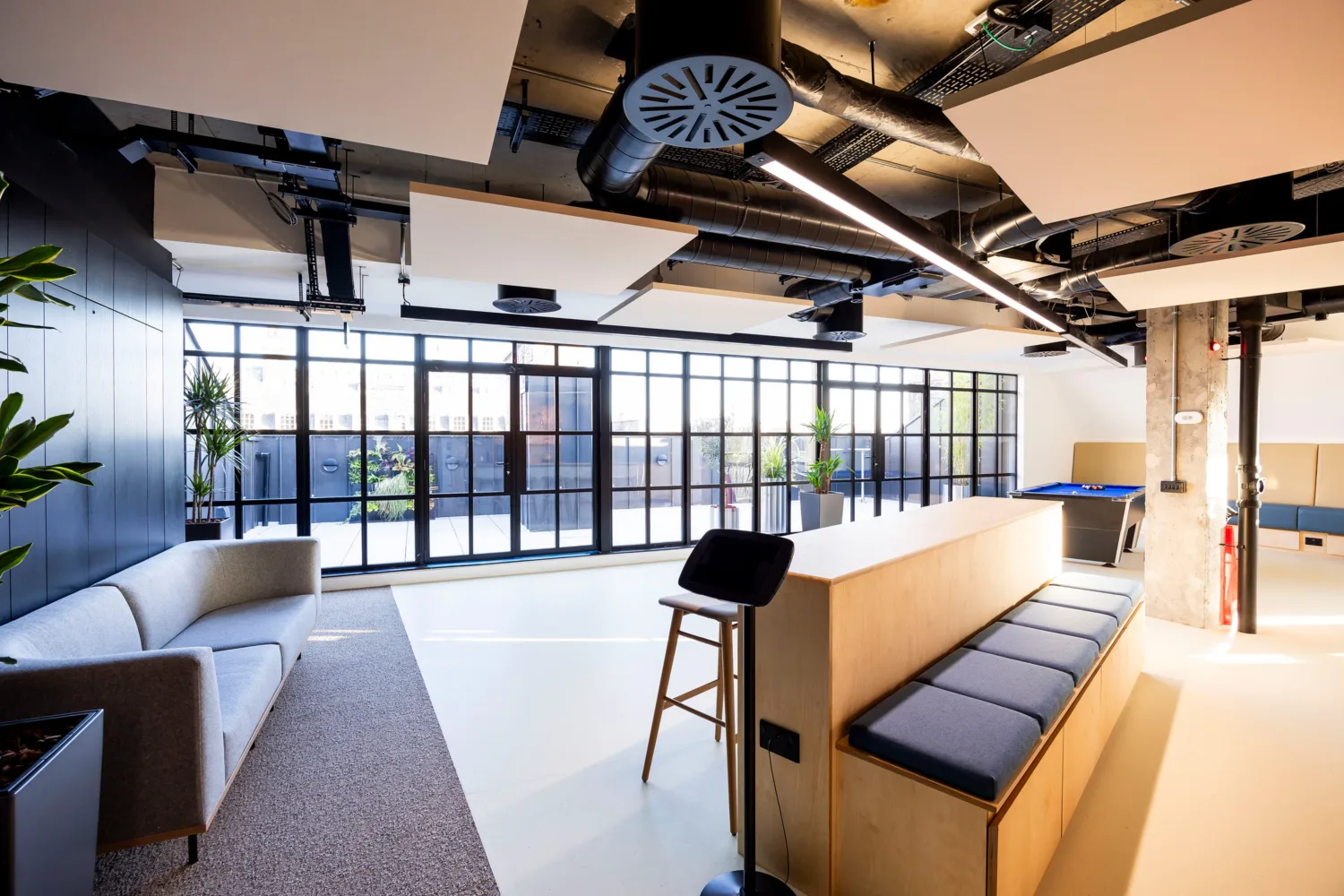 Modern office lounge with large windows, a long bench table, seating area, indoor plants, and exposed ceiling ducts. Natural light fills the space.