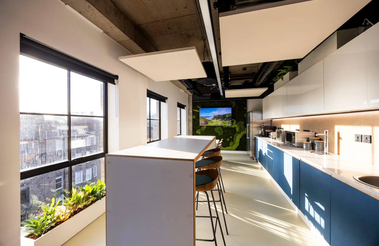 Modern office kitchen with high table, bar stools, large windows, plants, and built-in appliances. A screen displays a nature scene on the far wall. Natural light brightens the space.