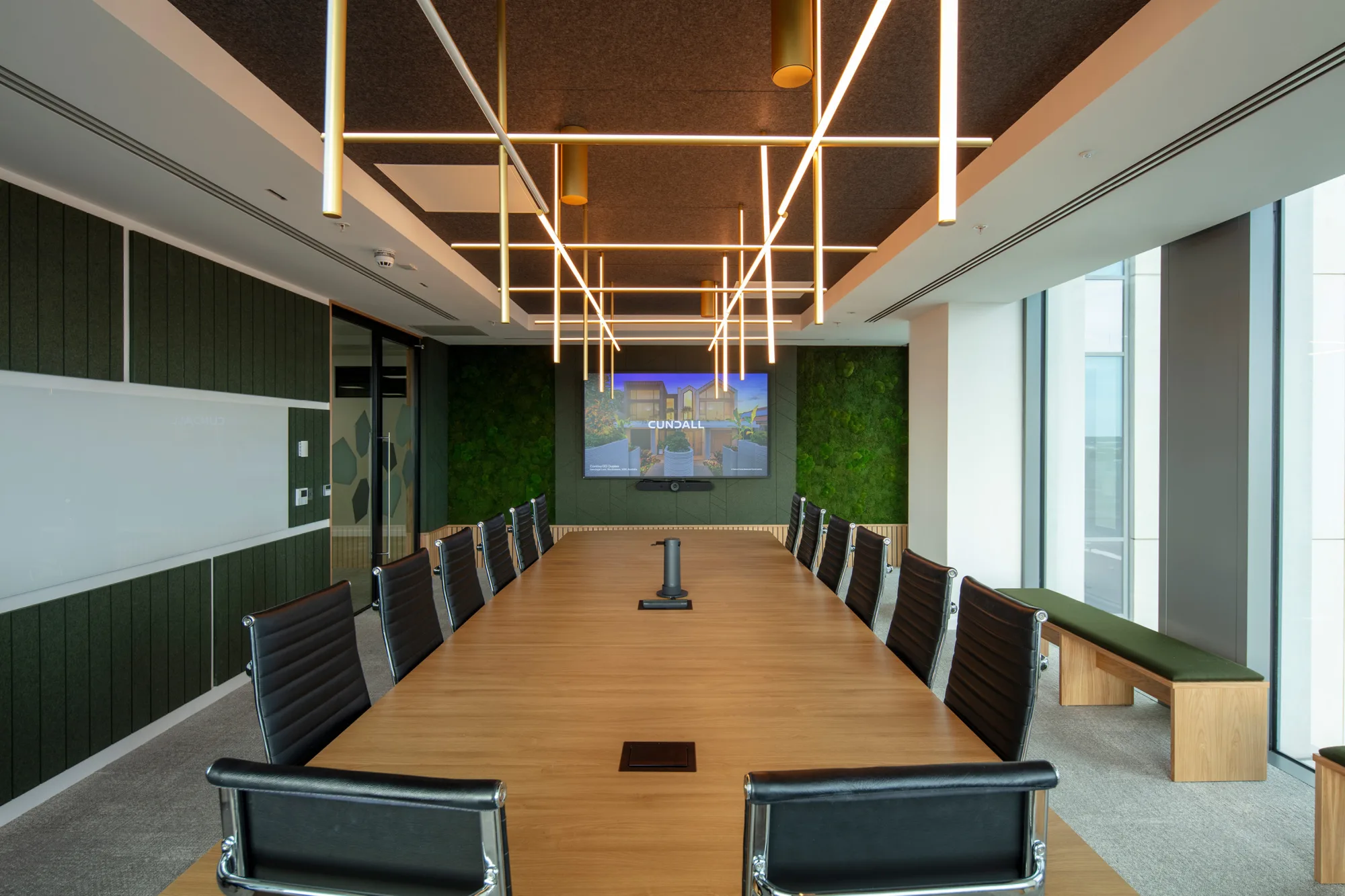 Conference room with a long wooden table surrounded by black chairs, a large screen displaying ‘Cundall’ on the far wall, green wall panels, whiteboard, and ceiling lights in a grid pattern.
