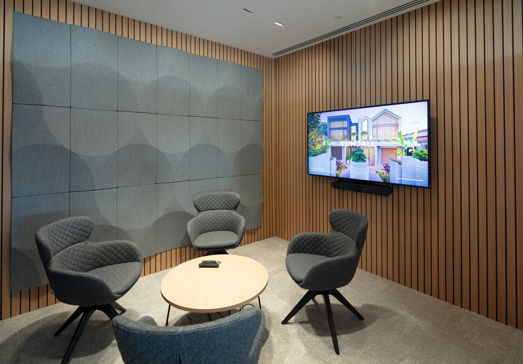 Small meeting area with four cushioned chairs around a round wooden table, remote control on the table, wood-paneled walls, gray acoustic panels, and a wall-mounted screen displaying a building image.