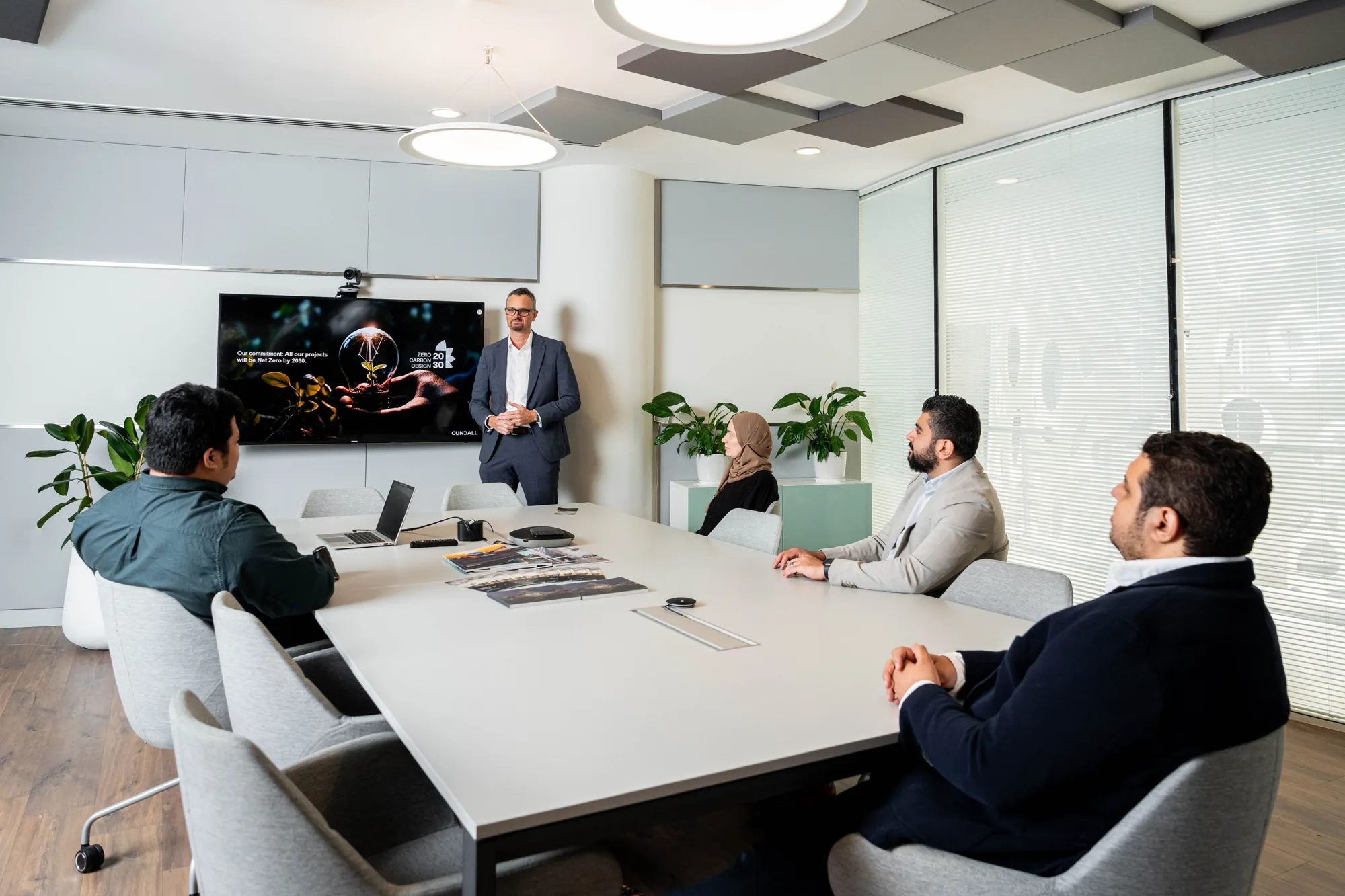 Meeting room with five people seated around a table facing a presenter standing beside a wall-mounted screen displaying a technical graphic. Laptops, documents, plants, glass partitions, and acoustic ceiling panels are visible.