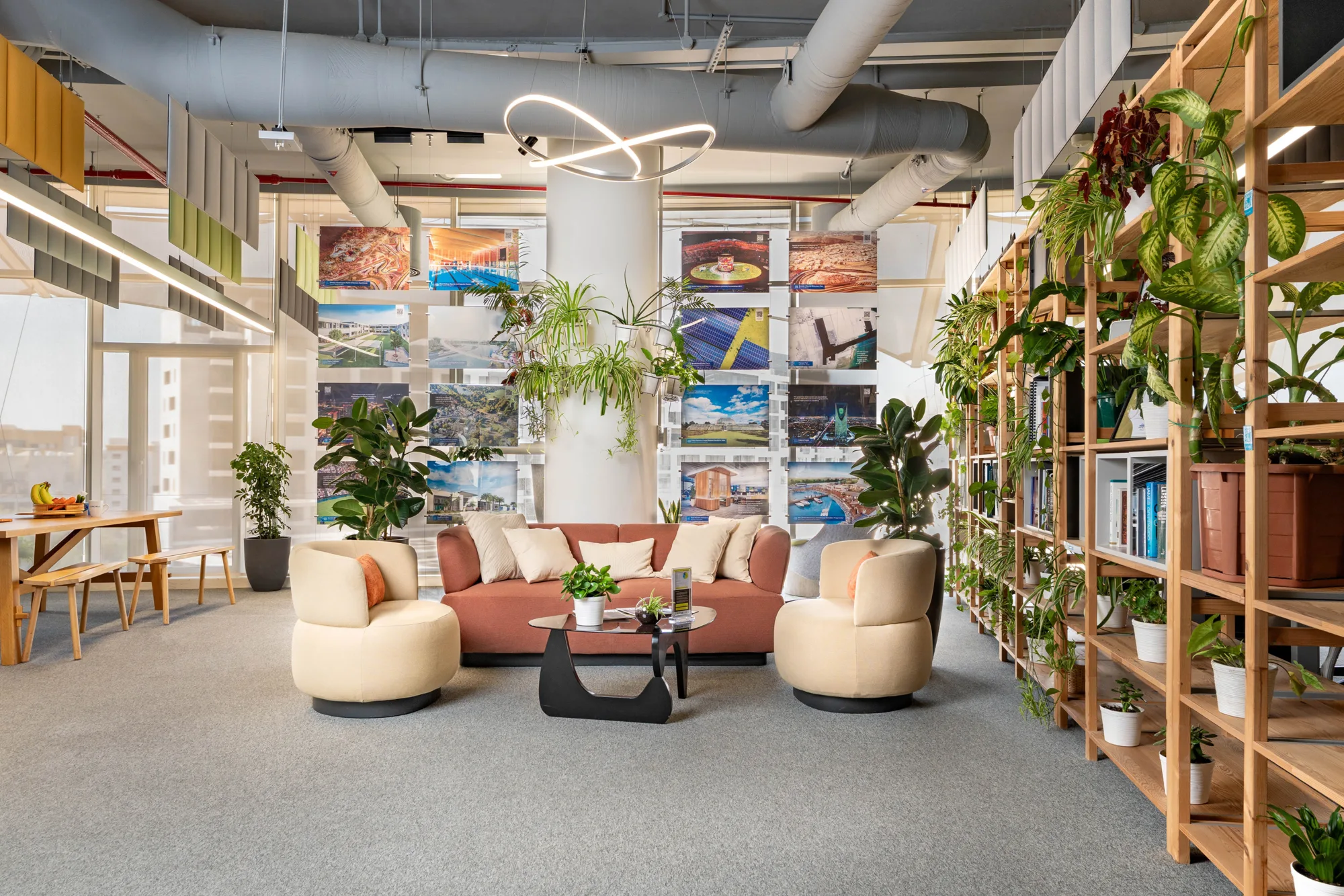 Open-plan office lounge with a central pink sofa, two rounded armchairs and a low table on a grey carpet. Wooden shelving filled with plants lines the right side, while large wall displays show project images. Exposed ceiling services and a feature light are visible above.