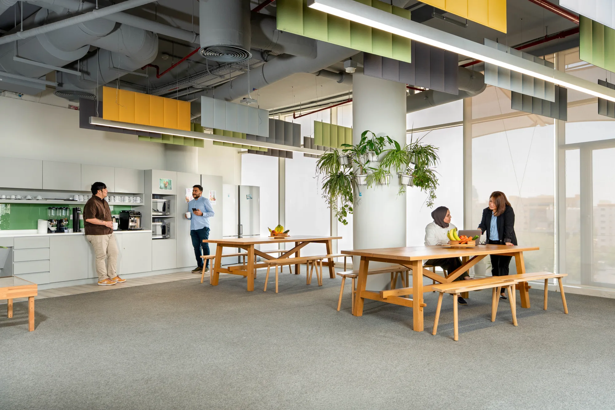 Open-plan office break area with wooden tables and benches, a kitchenette with cabinets and appliances on the left, and four people standing or seated. Large windows, indoor plants, grey carpet, and suspended acoustic panels are visible overhead.