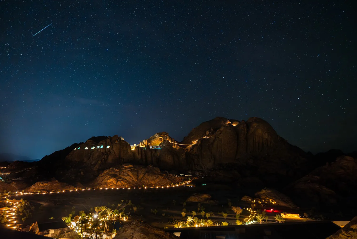 Star-filled night sky above rocky hills with illuminated pathways and structures across a desert landscape.