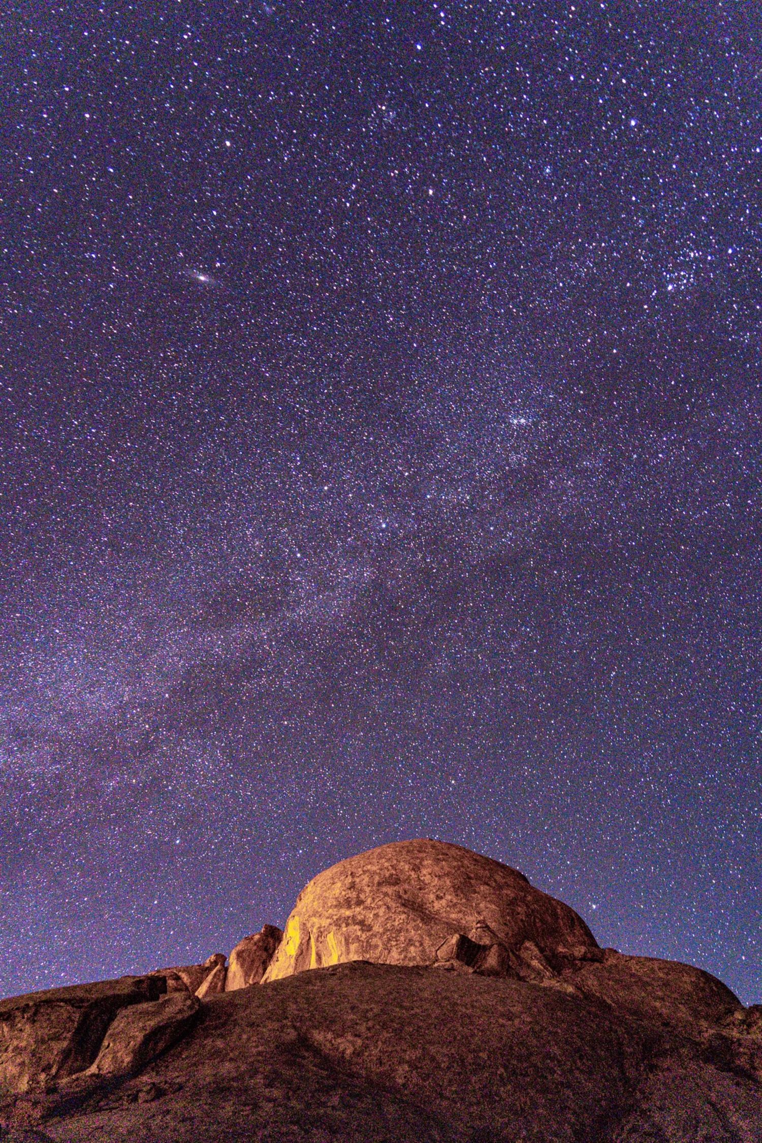 Star-filled night sky with the Milky Way visible above a large rock outcrop in a desert landscape.