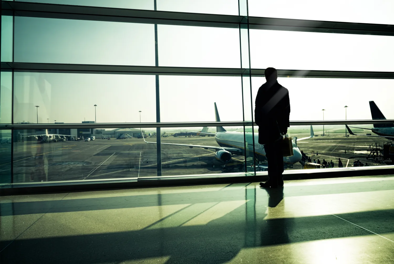 Silhouette of a person looking out of a window at an airport terminal outside which multiple parked planes are visible