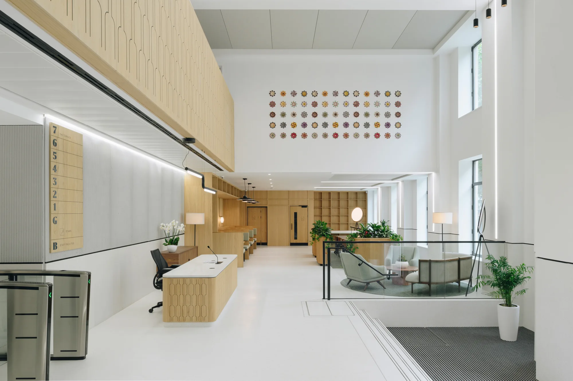 Modern office lobby with high ceiling and large windows. Left side has a reception desk, chair, computer, and turnstiles. Back wall has wooden cabinets and plants. Right side features seating, tables, lamps, and a wall with colorful circular patterns.