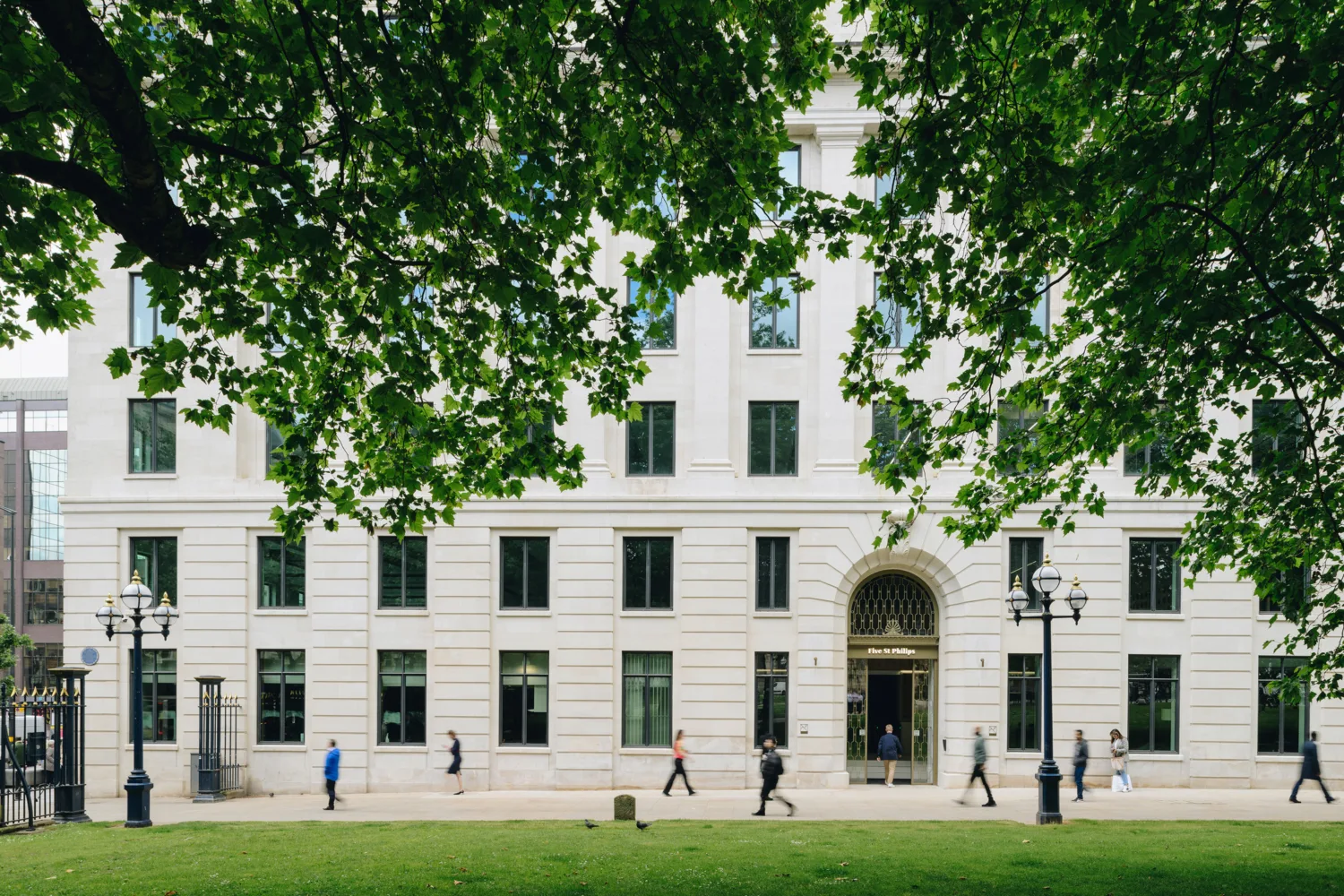 Large white stone building with multiple rectangular windows and an arched entrance, partially obscured by green tree branches. Several people walk on the sidewalk in front.