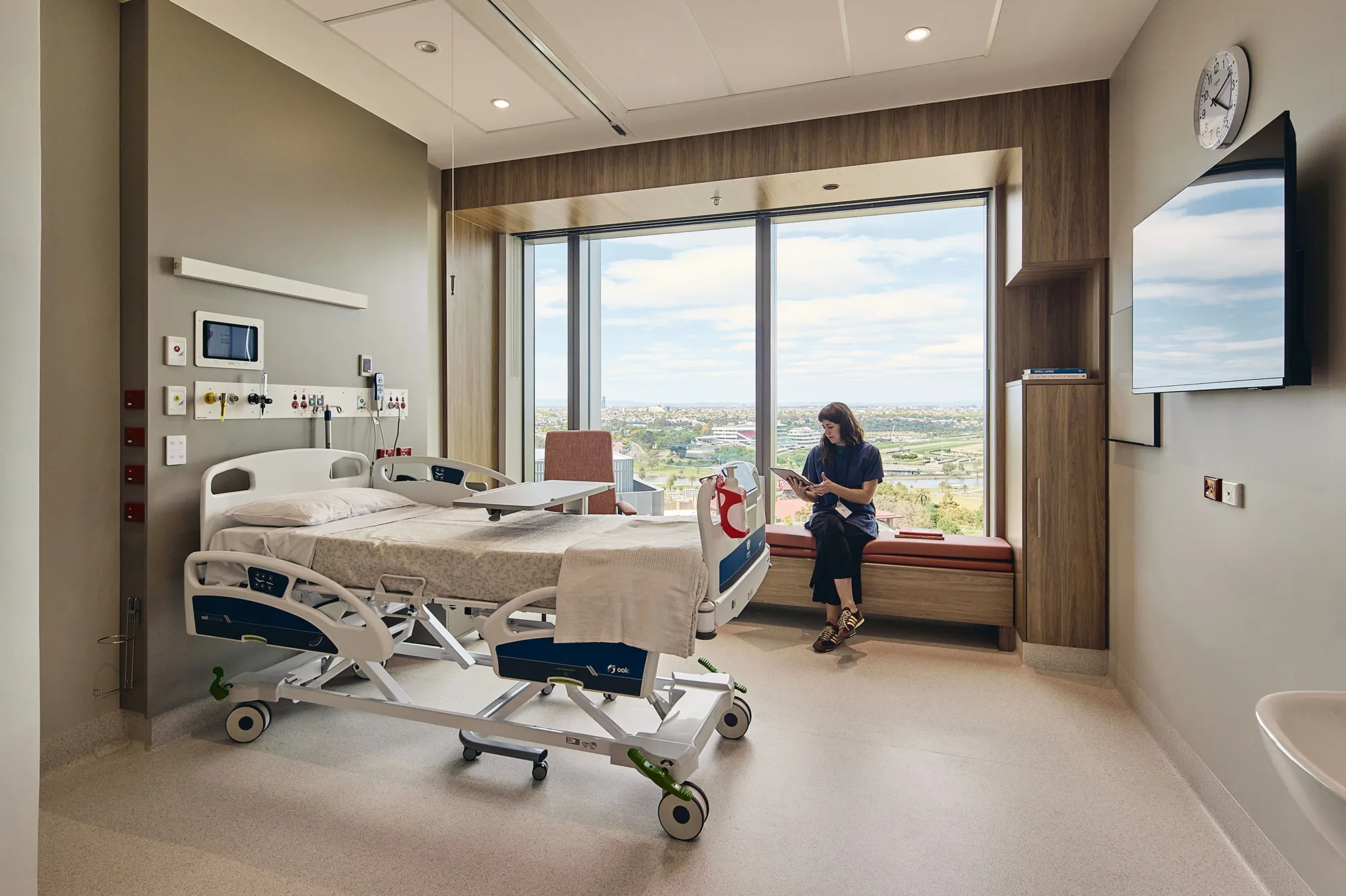 Patient room with an adjustable hospital bed, clinical wall services, large window seating area and city views.
