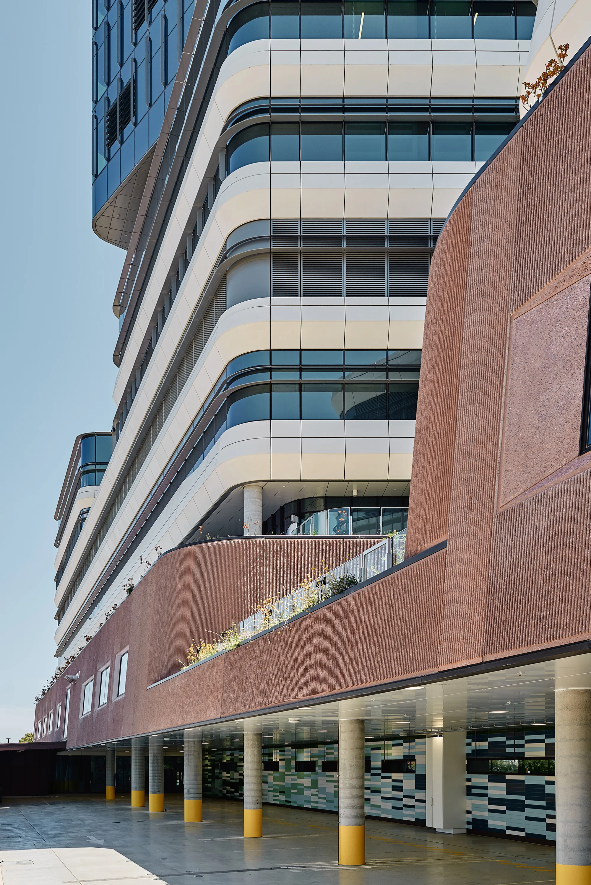 Close-up view of a multi-storey building façade with curved glazing, horizontal cladding bands, textured terracotta-coloured walls and covered ground-level walkways.