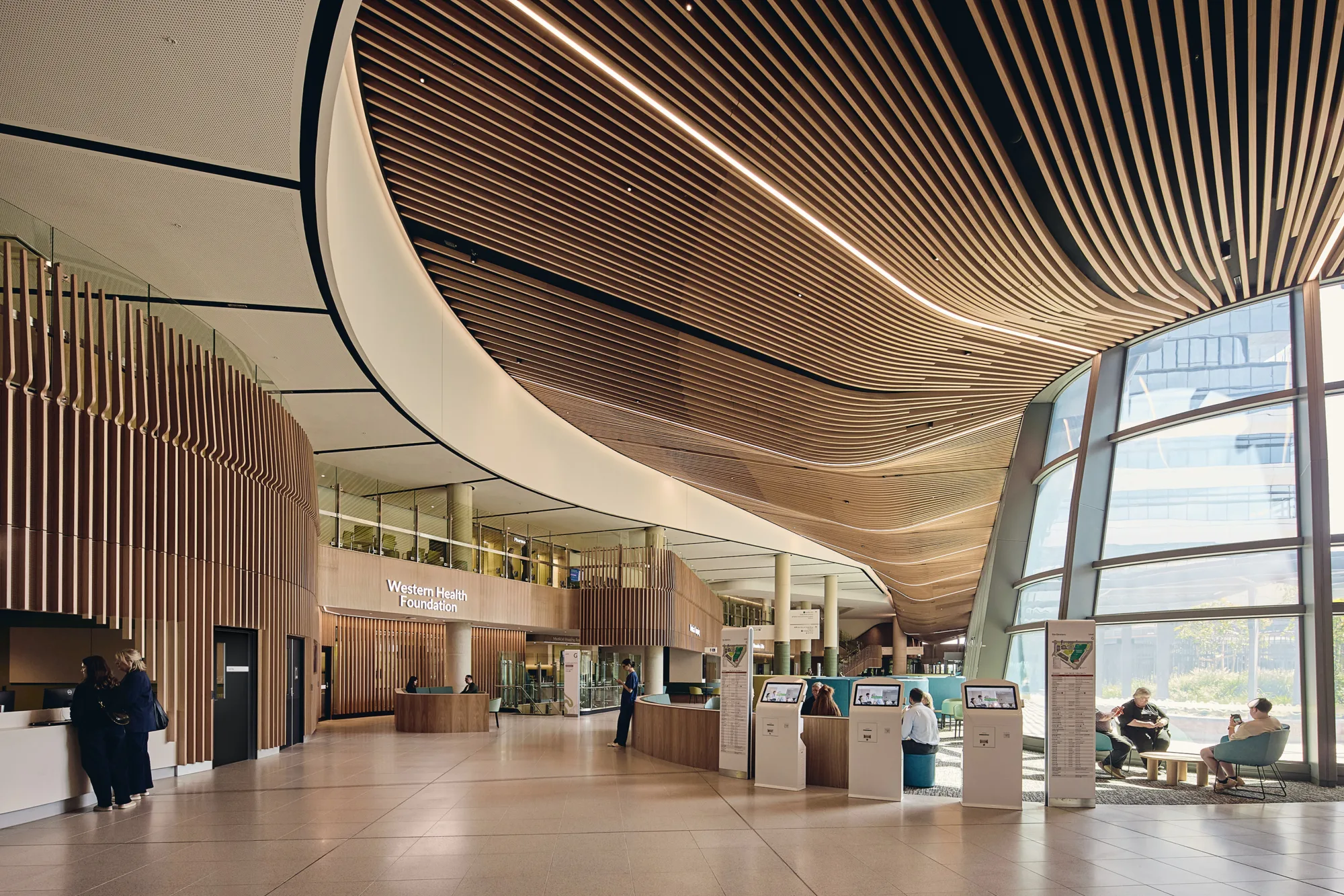 Hospital atrium with curved timber ceiling, digital wayfinding kiosks, seating areas and full-height glazed façade admitting natural daylight.