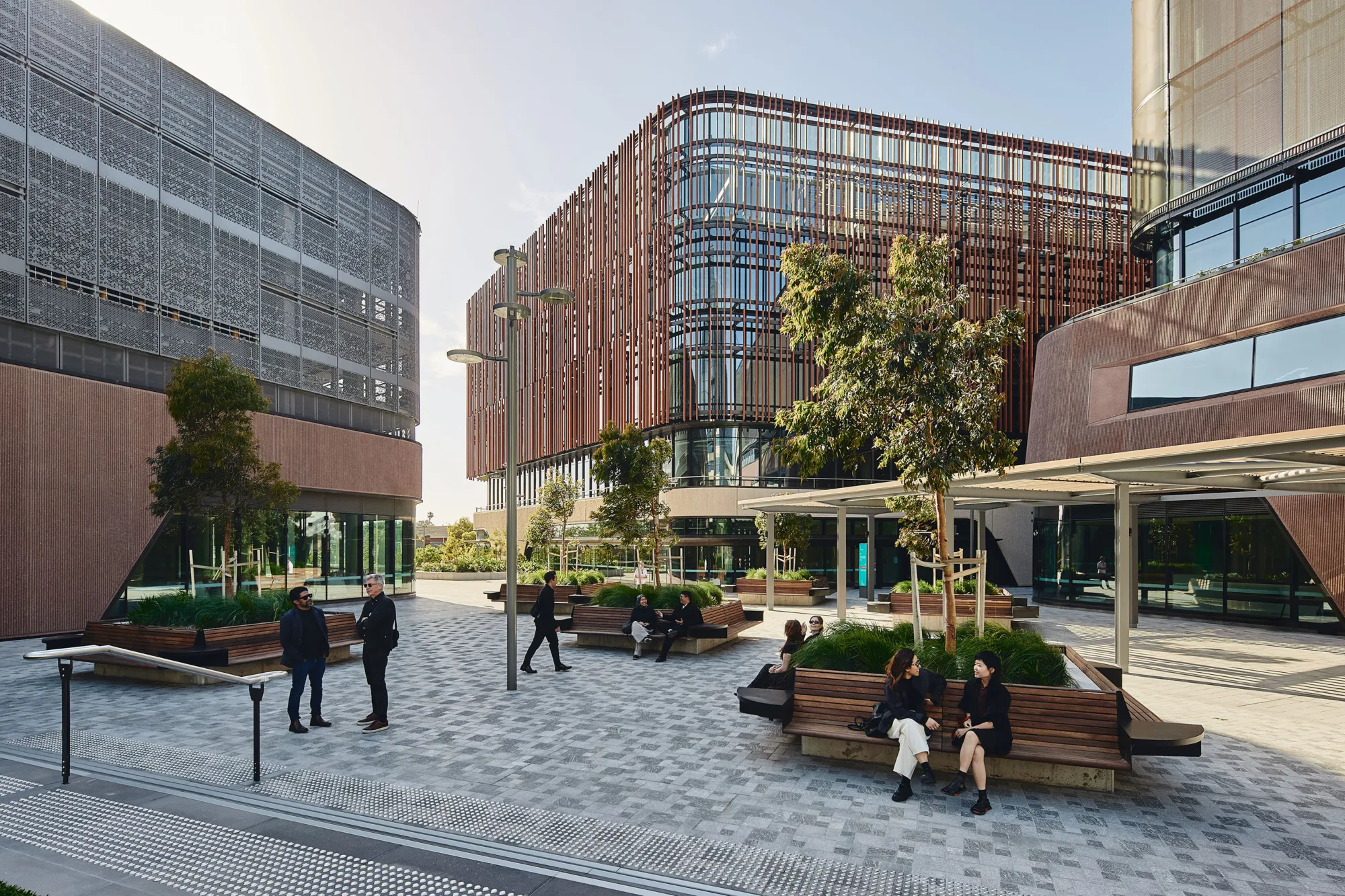 Landscaped outdoor courtyard between healthcare buildings with seating, trees, shaded walkways and glazed façades featuring vertical screening elements.