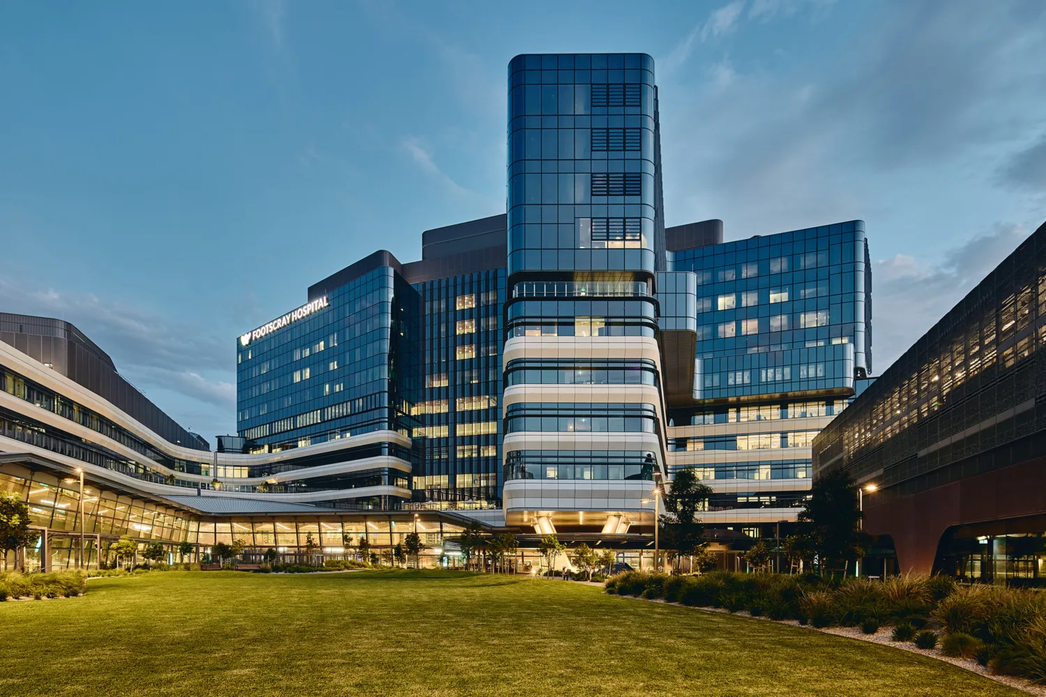 Modern healthcare building with glazed façades, illuminated windows and landscaped lawn viewed at dusk, with Footscray Hospital signage visible.