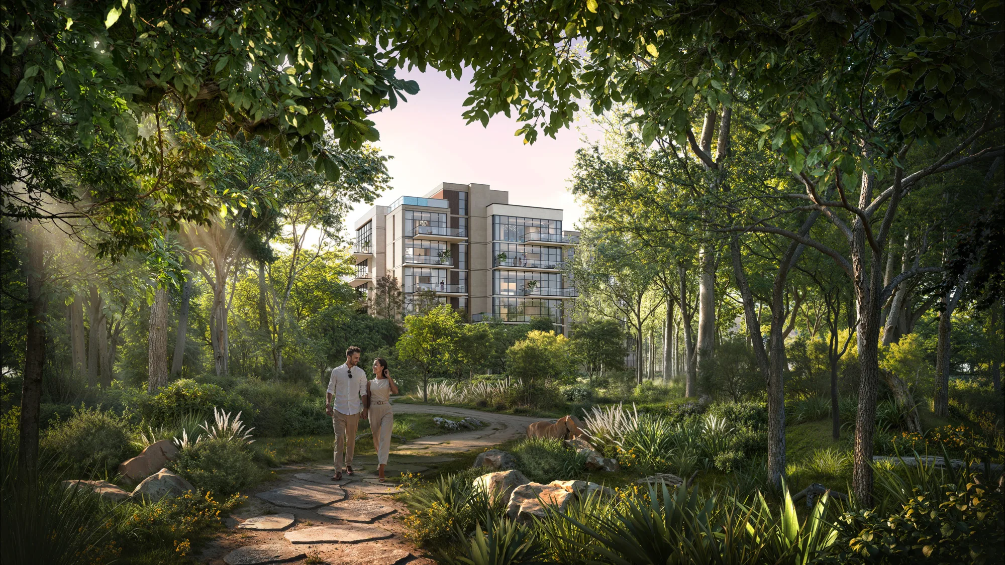Ground-level view of a winding stone path through dense trees and plants, with two people walking on the path, rocks and shrubs along the edges, and a modern multi-storey residential building visible through the foliage.
