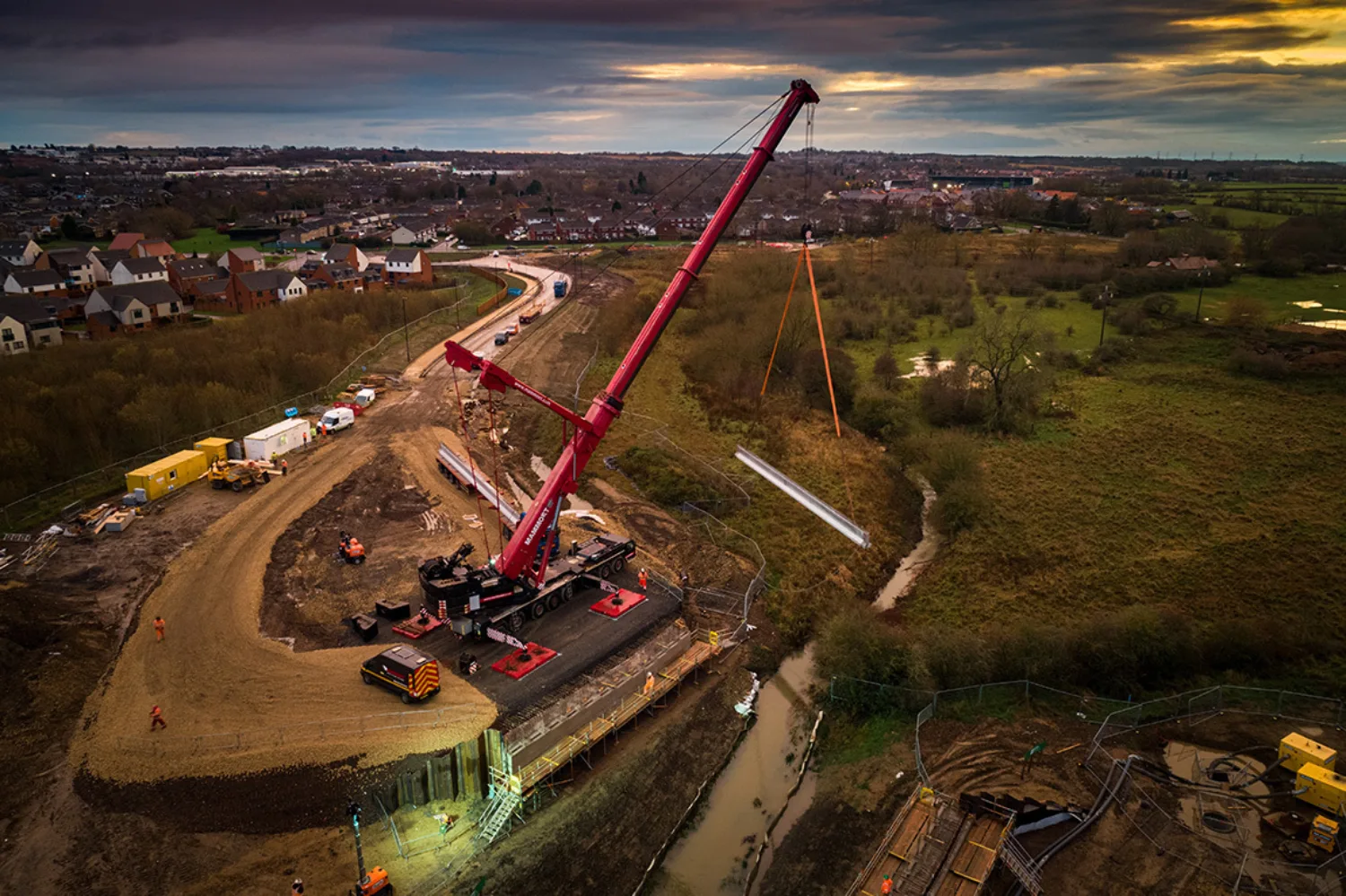 Red crane moving steel beam on a site surrounded by green vide, stream and housing developments