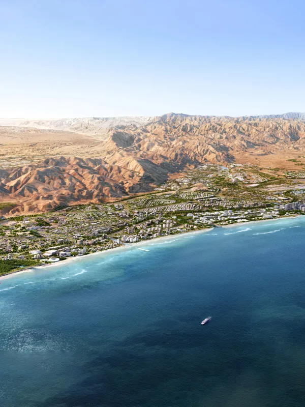 Aerial view of a coastal city with dense greenery, buildings, and roads along a curved shoreline. Clear blue water fills the foreground, and a wide expanse of rocky, beige mountains stretches across the background.