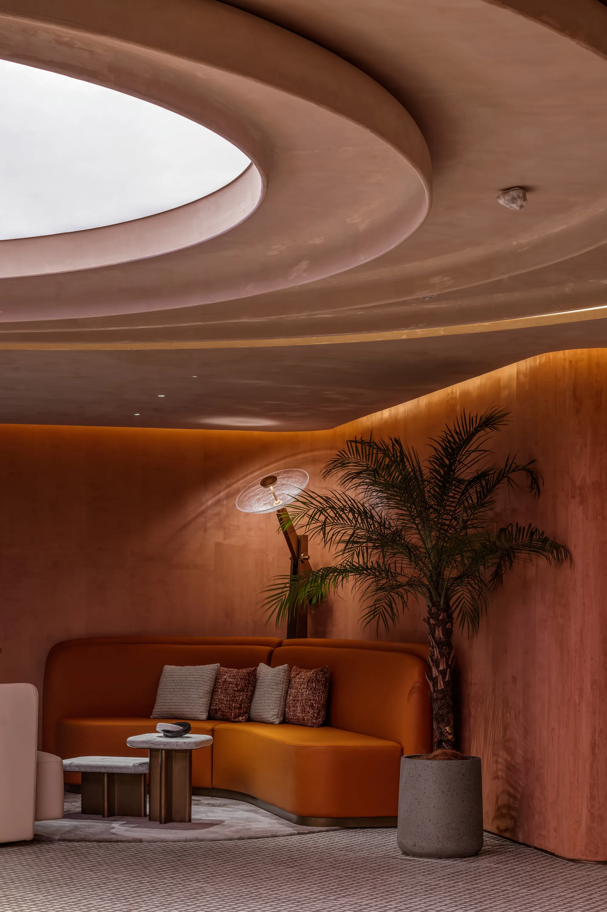 Curved copper-toned interior with a circular recessed ceiling opening and soft lighting. Below, a curved upholstered sofa with cushions surrounds small marble tables, a floor lamp, and a potted palm in a grey planter.